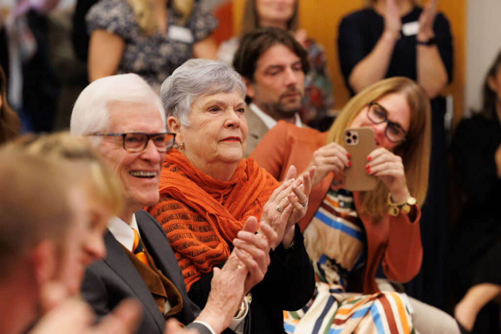 Attendees clapping at an event, including an older person in an orange scarf and a man next to her in a suit and orange tie.