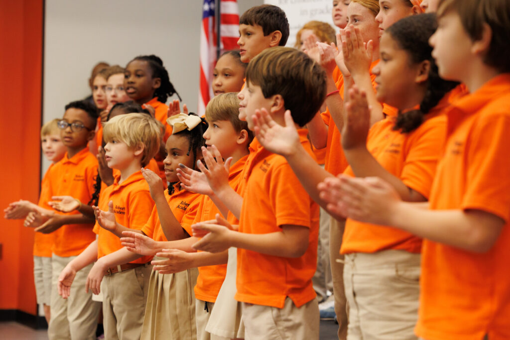 A group of children in orange shirts and khaki pants stand on stage, clapping their hands. An American flag is also visible in the background.