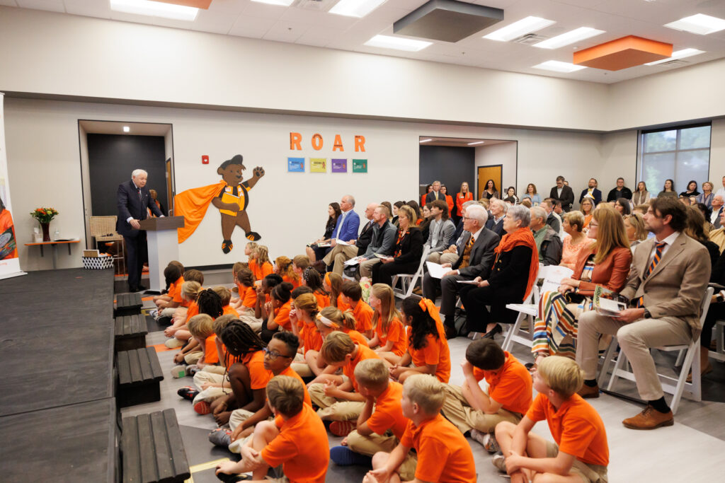 A speaker addressing an audience in an auditorium at an event, featuring a cub mascot on a wall labeled "ROAR". The audience is a mix of adults seated on chairs and children sitting on the floor, all attentively listening.