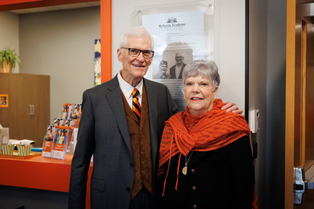 Two individuals standing in front of a plaque at an indoor event, smiling at the camera.