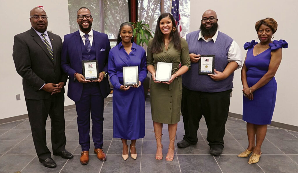 Six people stand in a row. The four people in the middle hold plaques.