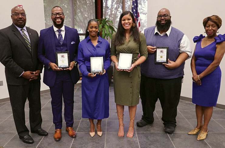 Six people stand in a row. The four people in the middle hold plaques.