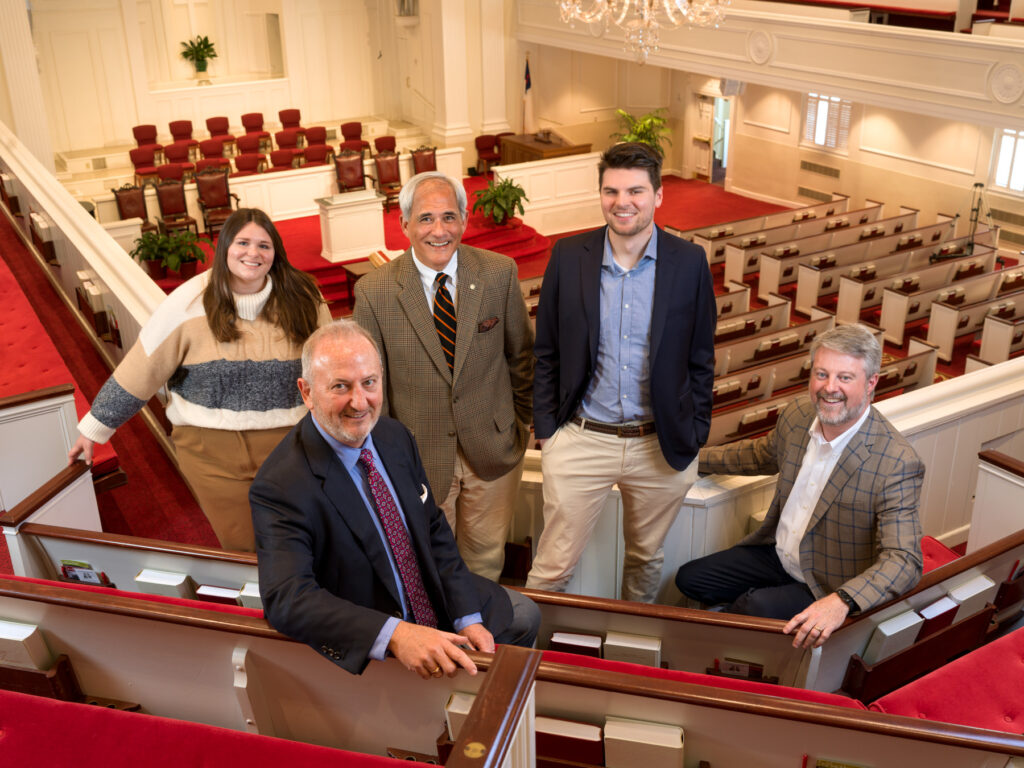 Five individuals posing for a photo in a church sanctuary with rows of red pews and a white interior.