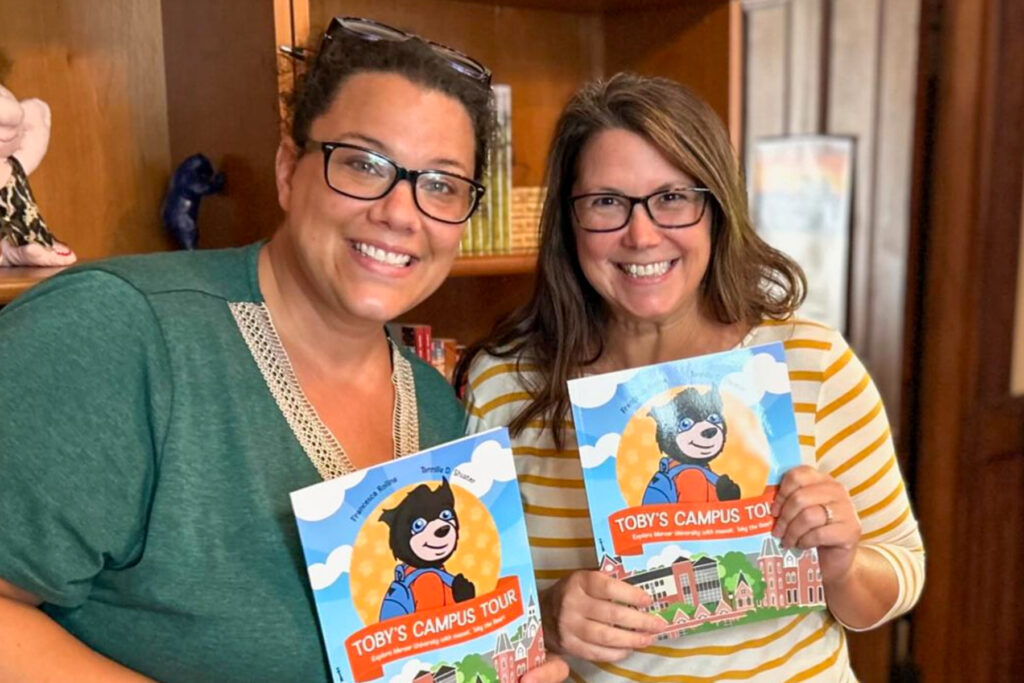 Two individuals smiling and holding copies of the children's book "Toby's Campus Tour" inside a room.