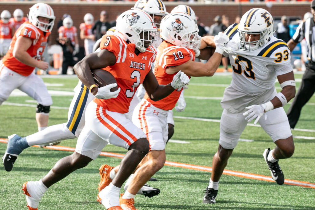 Mercer and ETSU football players compete on a field, with No. 3 from Mercer carrying the ball.