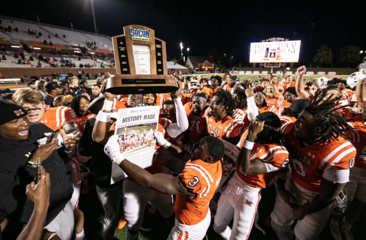 Mercer football team celebrates on the field with a trophy and newspaper headlined History Made.