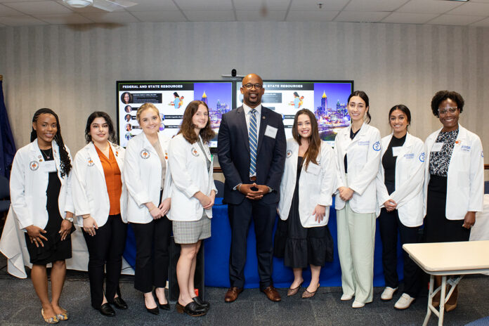 Group of eight medical students wearing white lab coats and one person in a suit standing together in a room.