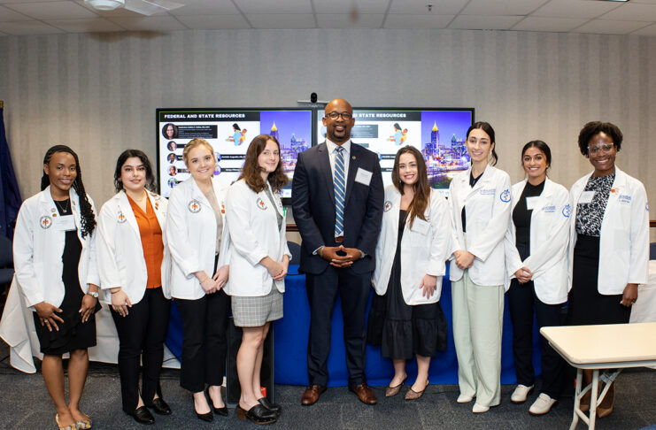 Group of eight medical students wearing white lab coats and one person in a suit standing together in a room.