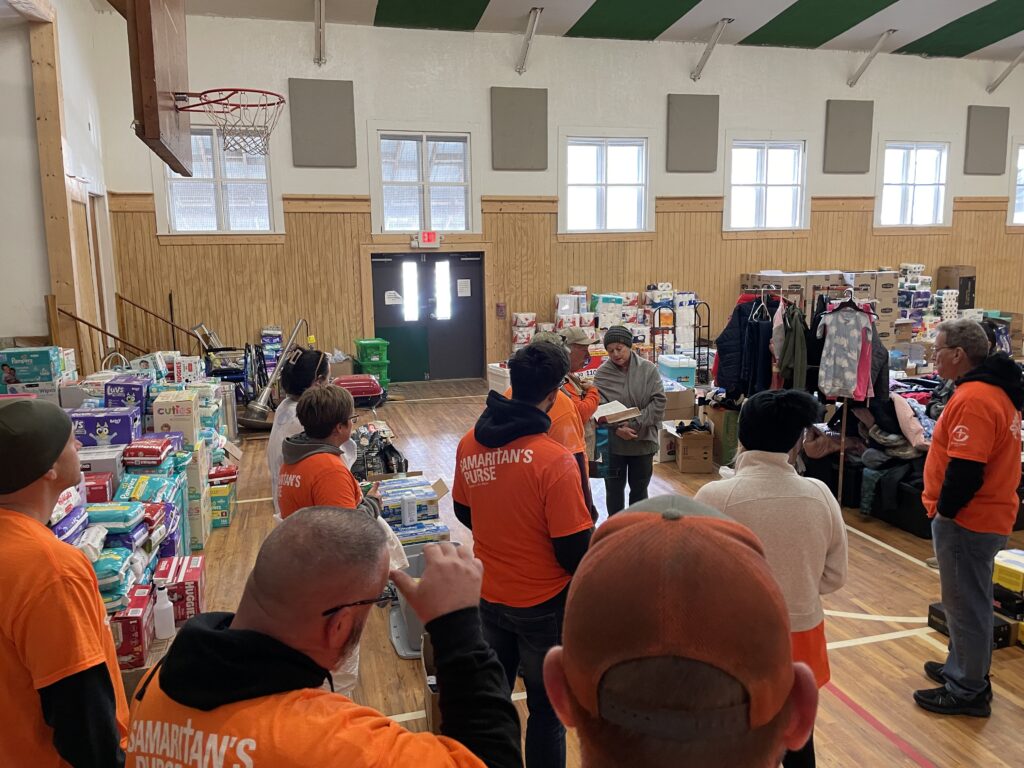 A group of volunteers in orange shirts gathers in a gymnasium filled with donated supplies.