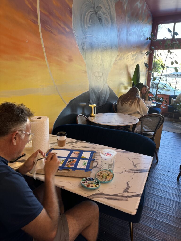 Patrons sit at tables at a coffee shop, with a man painting in the foreground.