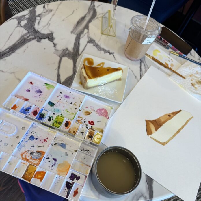 A cafe table with watercolor paints, water, a cheesecake, and a piece of paper with a painting of the cheesecake.