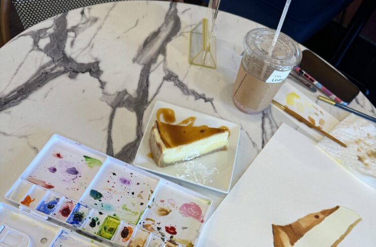 A cafe table with watercolor paints, water, a cheesecake, and a piece of paper with a painting of the cheesecake.