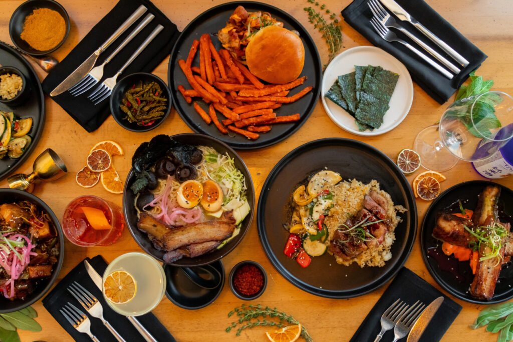 An overhead view of a table with various dishes, including ramen bowls, a burger and fries, and steak on top of rice.