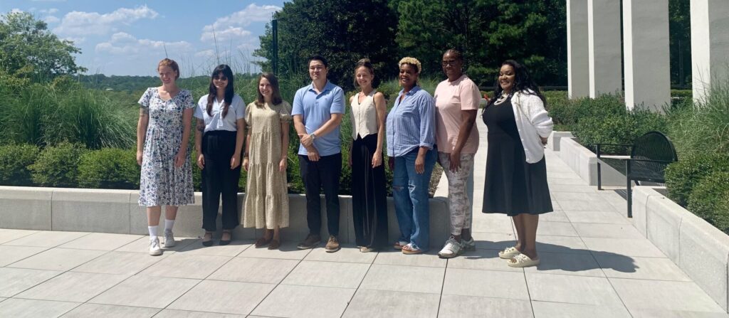 A group of eight people stand outside, smiling and posing in front of greenery and white columns.