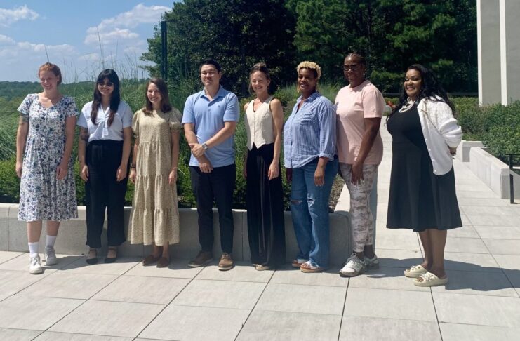 A group of eight people stand outside, smiling and posing in front of greenery and white columns.