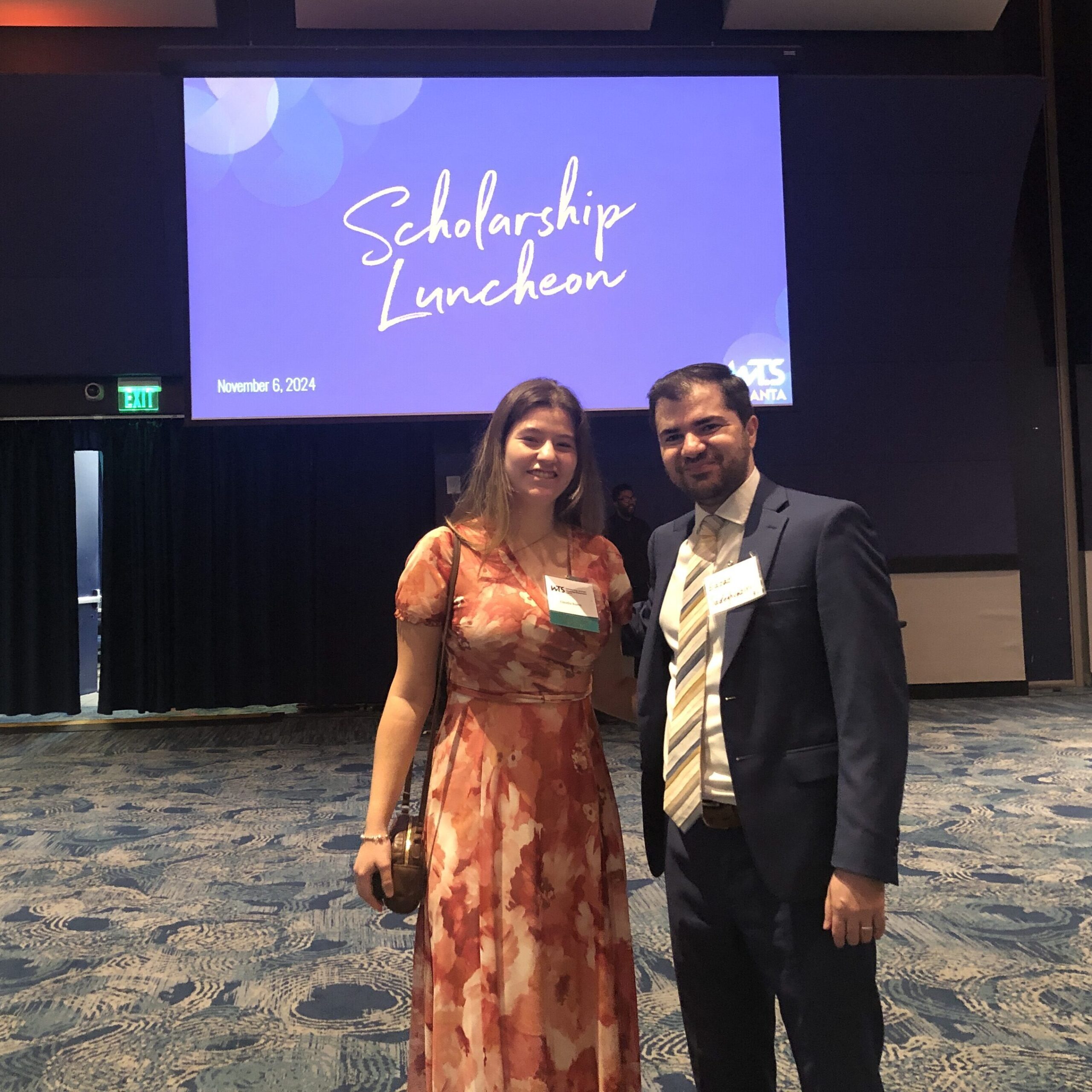 Two people standing together at a scholarship luncheon event with a presentation screen in the background.