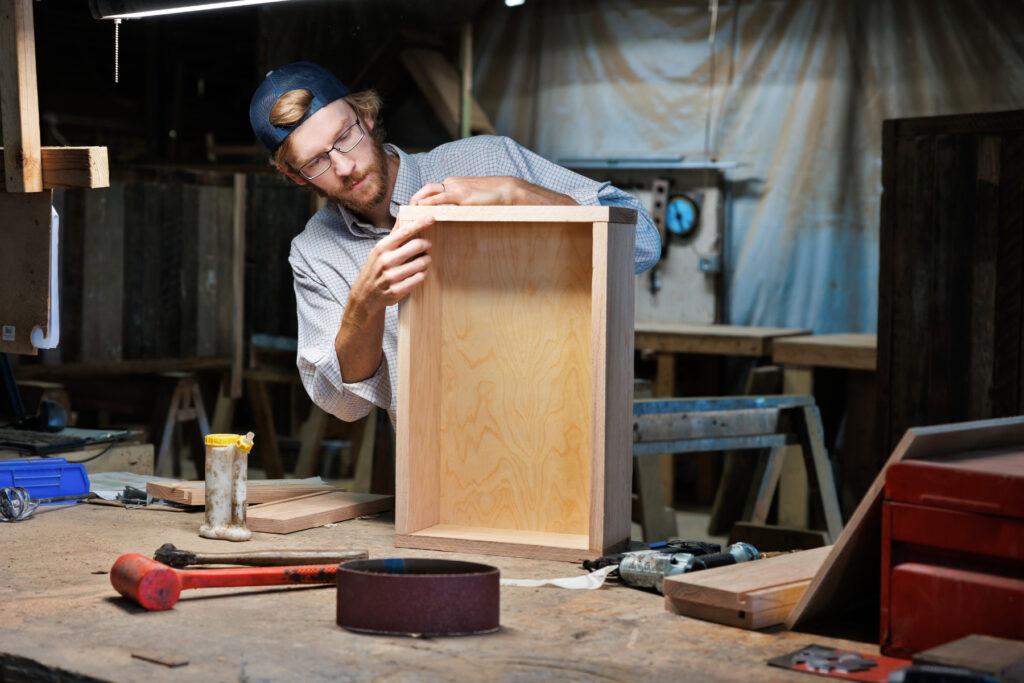 Andrew Eck assembles a wooden cabinet in a workshop filled with tools and wood materials.