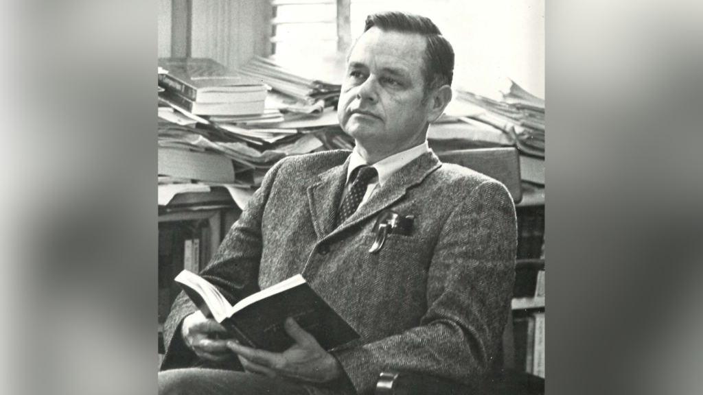 Dr. Benjamin Woodward Griffith Jr. sits in a chair holding an open book, with stacks of books in the background.