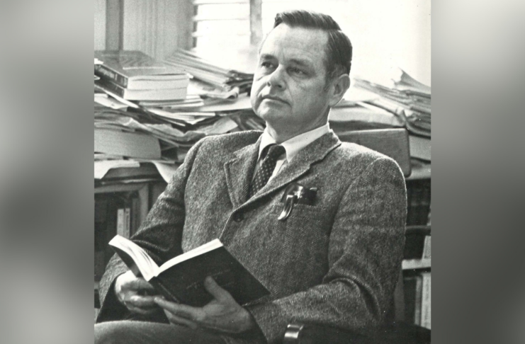 Dr. Benjamin Woodward Griffith Jr. sits in a chair holding an open book, with stacks of books in the background.