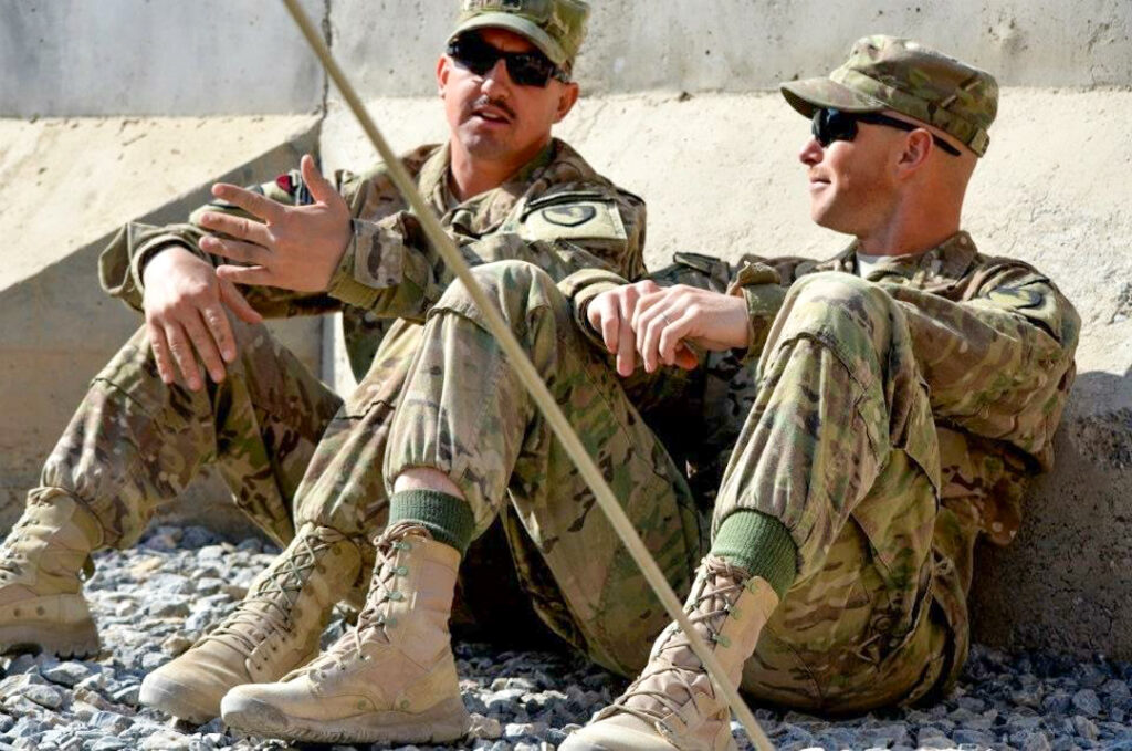 Craig Mattoon and another soldiers sit on the ground, conversing near a concrete wall.