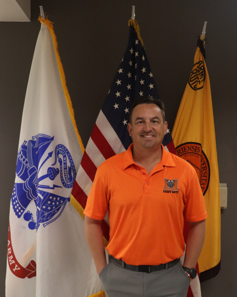 Craig Mattoon stands in front of a U.S. Army flag, an American flag, and an Army ROTC flag.