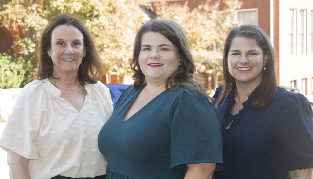 Three women standing outdoors, smiling, with buildings and trees in the background.