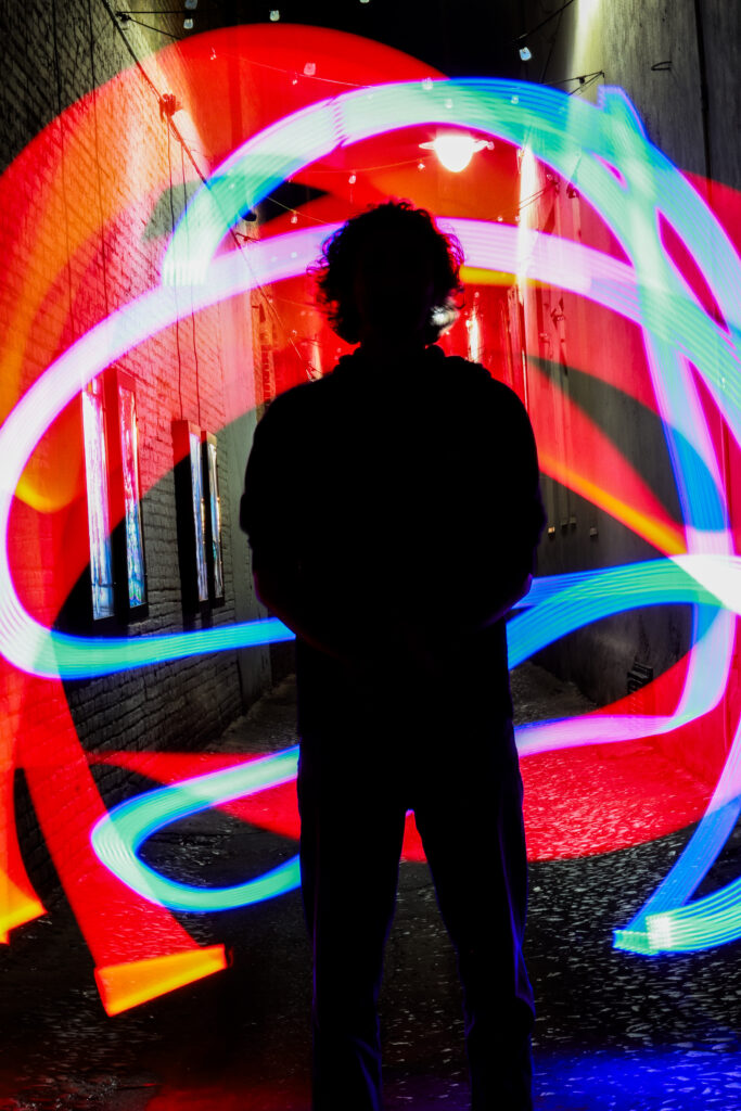 A silhouette of a person is in front of ribbons of colored light in an alleyway.