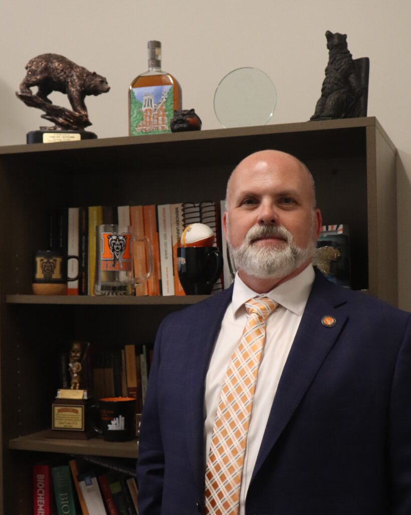 Garland Crawford, wearing a navy blue suit and orange checkered tie, stands in front of a book shelf.