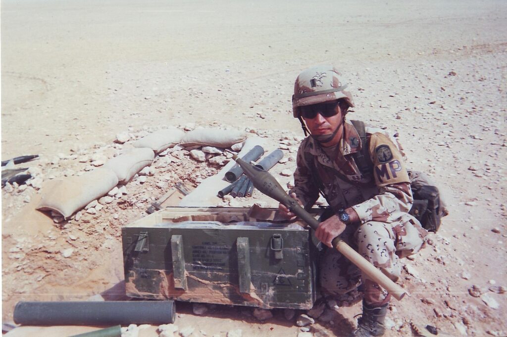 Michael Junod wearing Army fatigues and crouched down next to a an Army green chest.