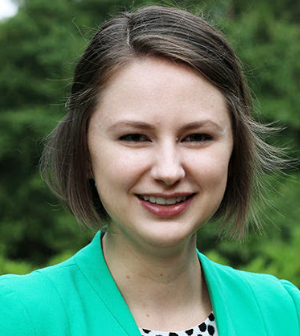 Portrait of a smiling Leah Panther wearing a green jacket and a polka-dot top, standing in front of greenery.