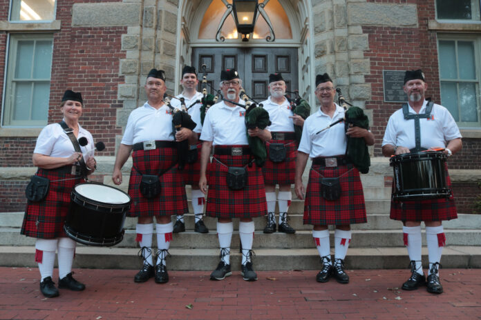 Group of seven people in traditional Scottish attire, including kilts, standing on steps with bagpipes and drums.