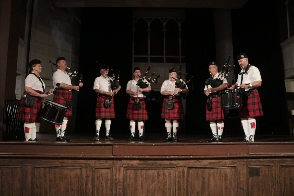 Members of a pipe band, dressed in tartan kilts and white shirts, perform on stage with bagpipes and drums.