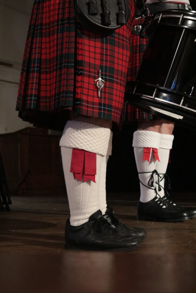 Person in traditional Scottish attire with a tartan kilt and white socks with red flashes, holding a drum.