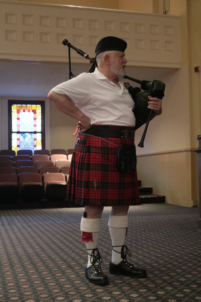 Person in traditional Scottish attire, including a tartan kilt, holding the bagpipes in a room with stained glass windows.