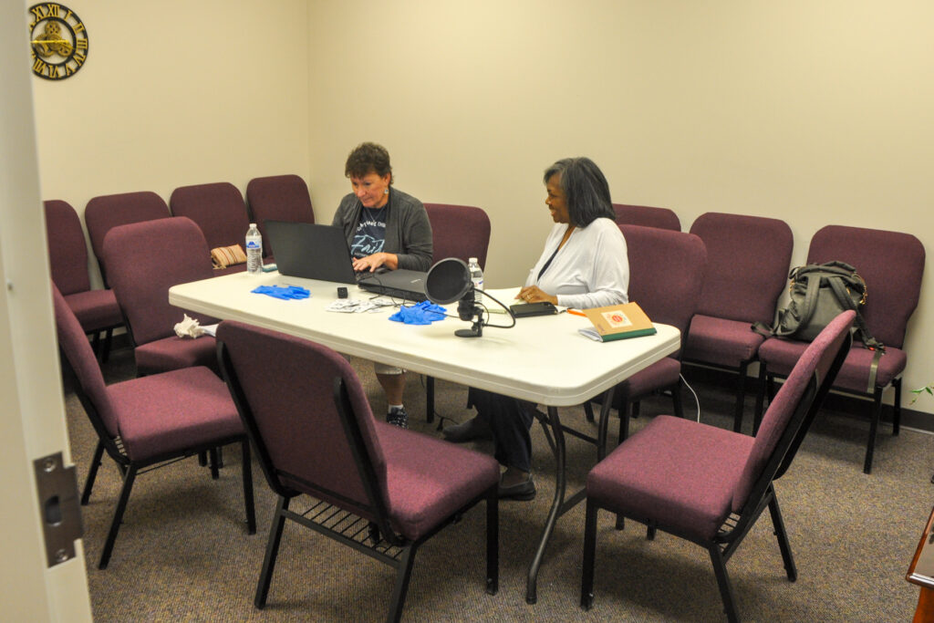 Two individuals are seated at a table working on a laptop in a conference room with several empty chairs around them.