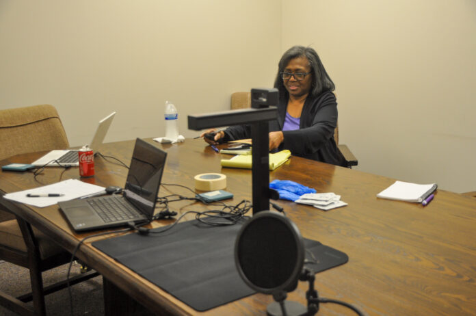 Person sitting at a conference table with multiple electronic devices including laptops and a microphone in a meeting room setting.