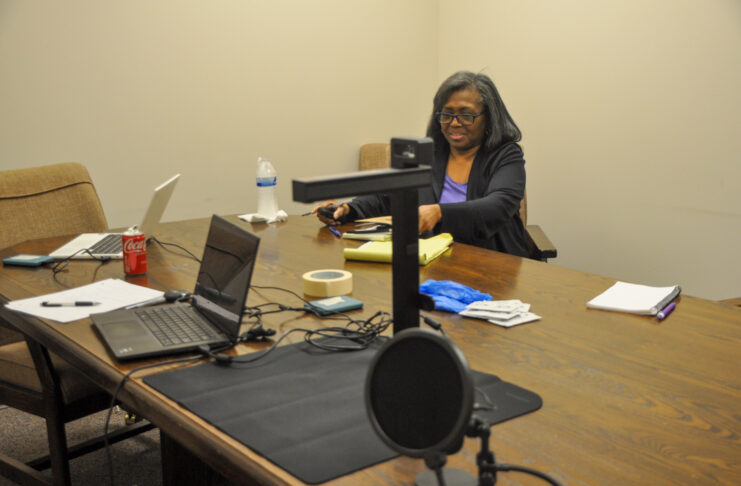 Person sitting at a conference table with multiple electronic devices including laptops and a microphone in a meeting room setting.