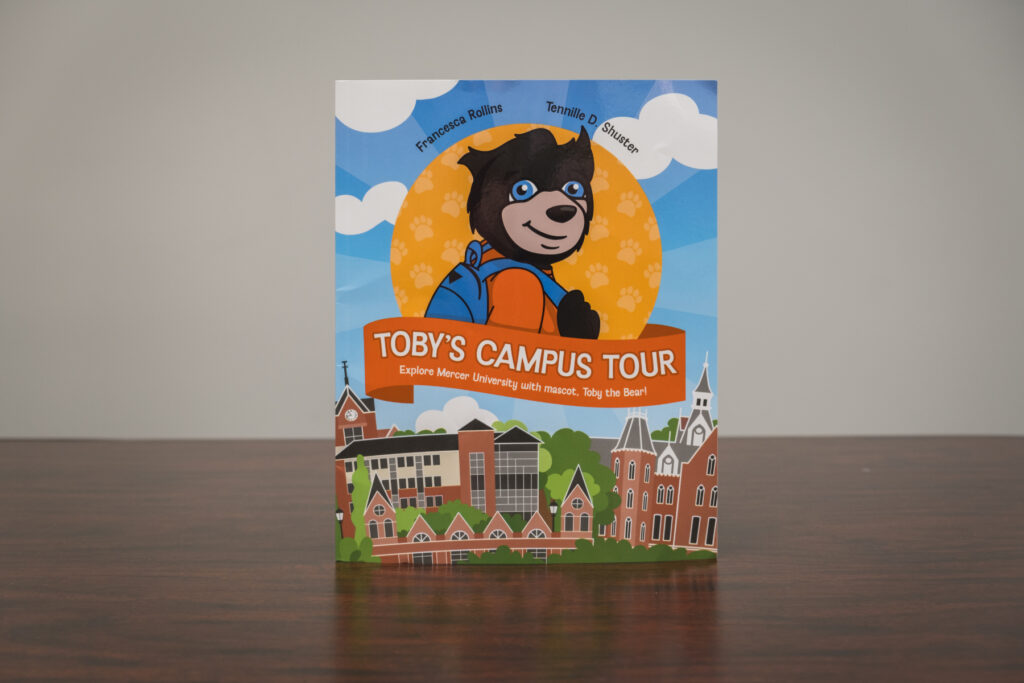 "Toby's Campus Tour" book cover featuring a cartoon Toby wearing a backpack against a Mercer campus backdrop.