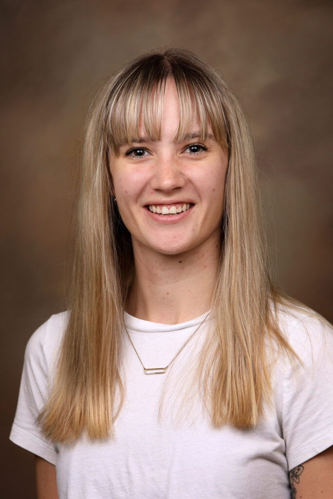 A person with long straight blonde hair and bangs smiles at the camera, wearing a white shirt and a necklace.