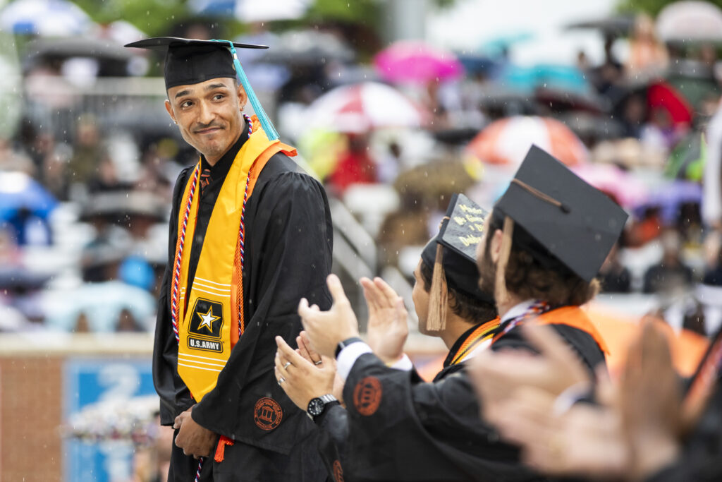 Graduating student in a cap and gown stands amid rain, wearing a U.S. Army stole, as others applaud.