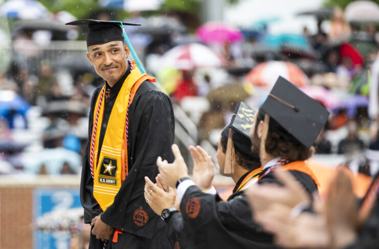 Graduating student in a cap and gown stands amid rain, wearing a U.S. Army stole, as others applaud.