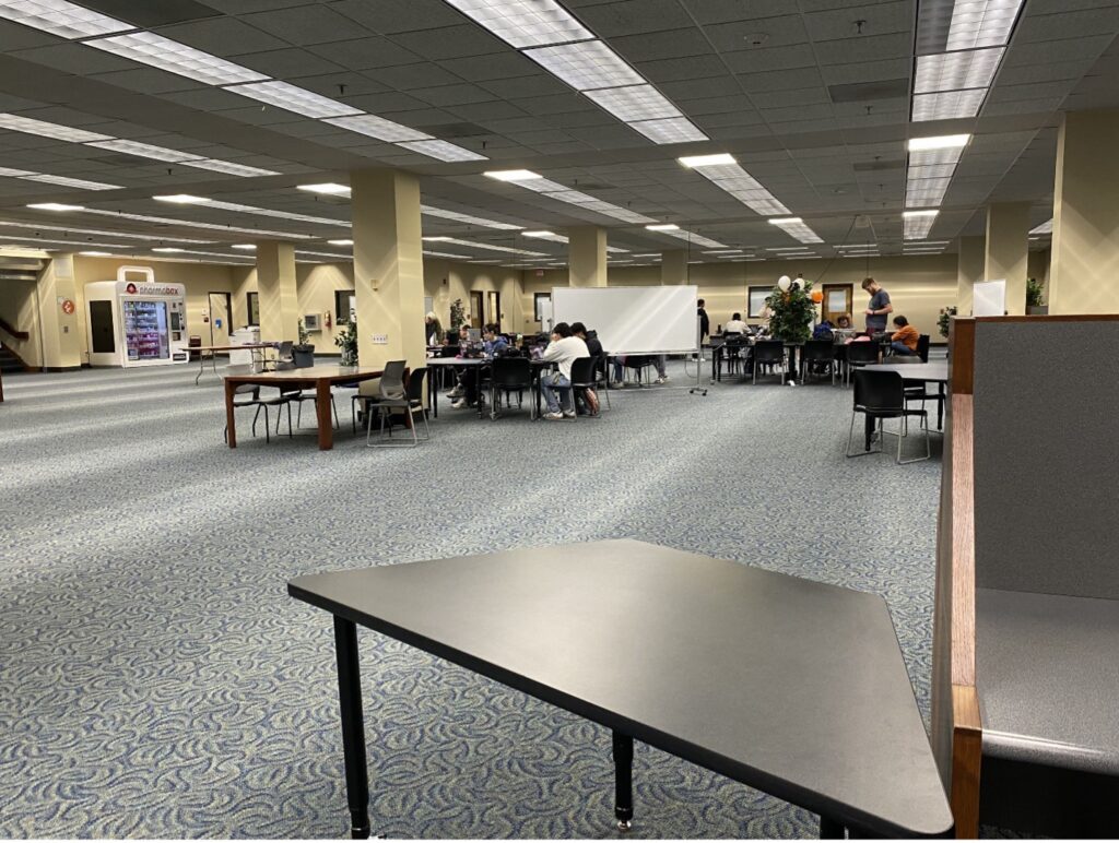 Wide-angle view of a spacious library with tables, chairs, and people studying in groups.