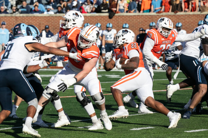 A college football game between Mercer and Rhode Island with Mercer players blocking and running while Rhode Island players attempt to tackle.