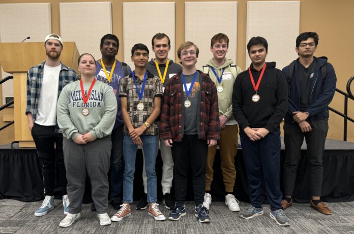 A group of nine people standing indoors, some wearing medals around their necks.