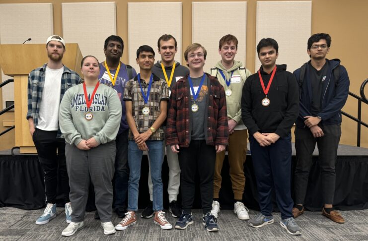 A group of nine people standing indoors, some wearing medals around their necks.