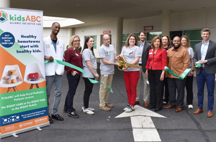 A group of people stand outside cutting a green ribbon next to a sign that reads kids ABC Alliance for Better Care.