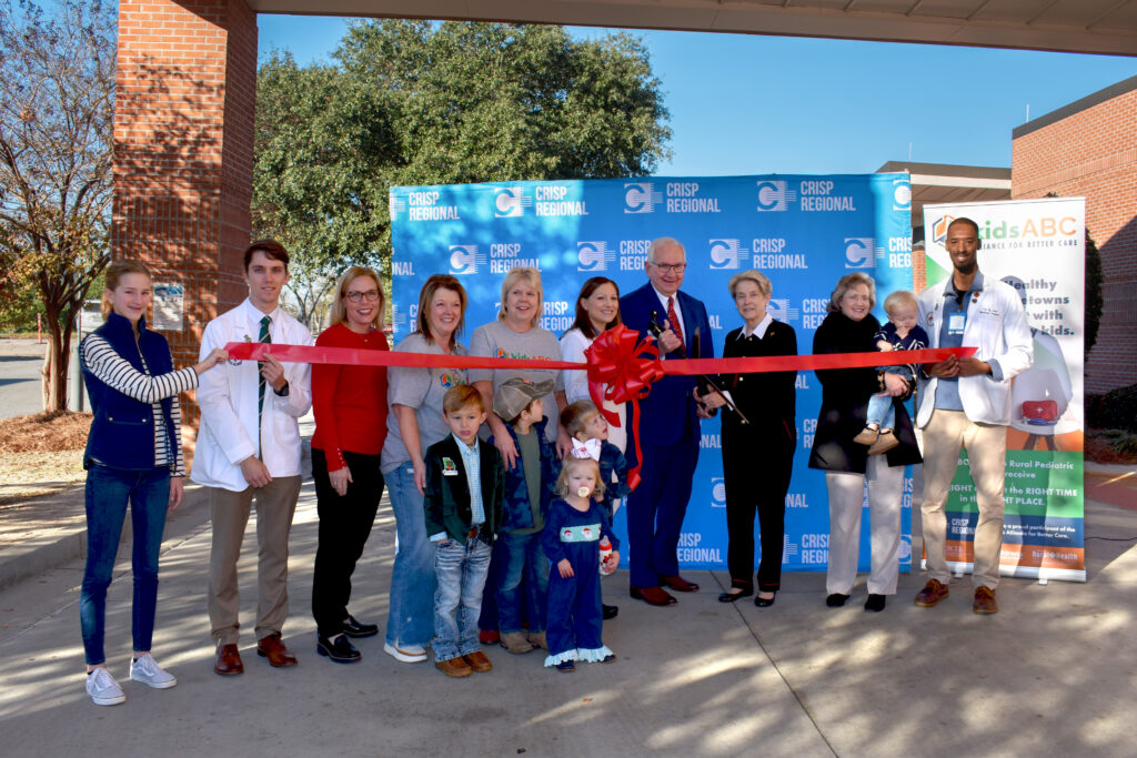 A group of people participate in a ribbon-cutting ceremony outside, with a red ribbon and a Crisp Regional backdrop.