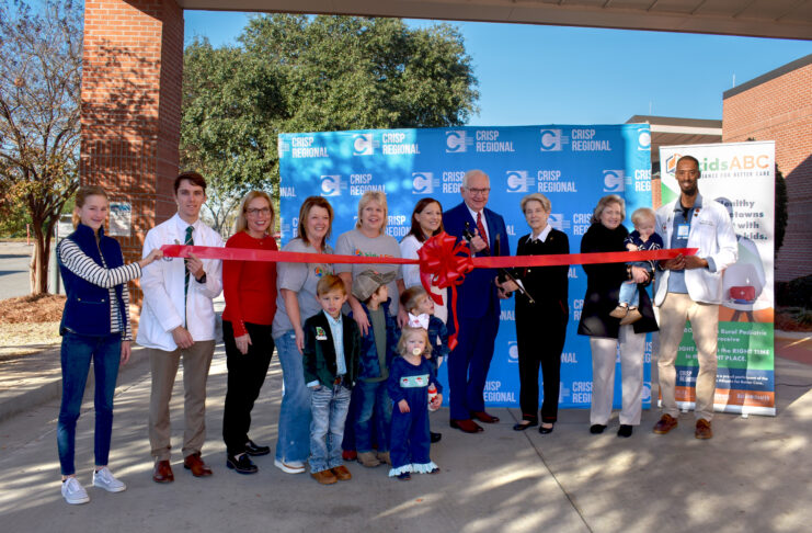 A group of people participate in a ribbon-cutting ceremony outside, with a red ribbon and a Crisp Regional backdrop.