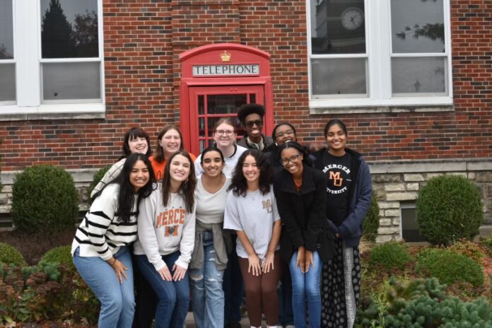 A group of people smile in front of a red telephone booth against a brick building.