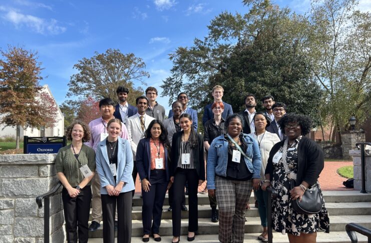 A diverse group of people stands on outdoor steps, smiling, with trees and a brick path in the background.